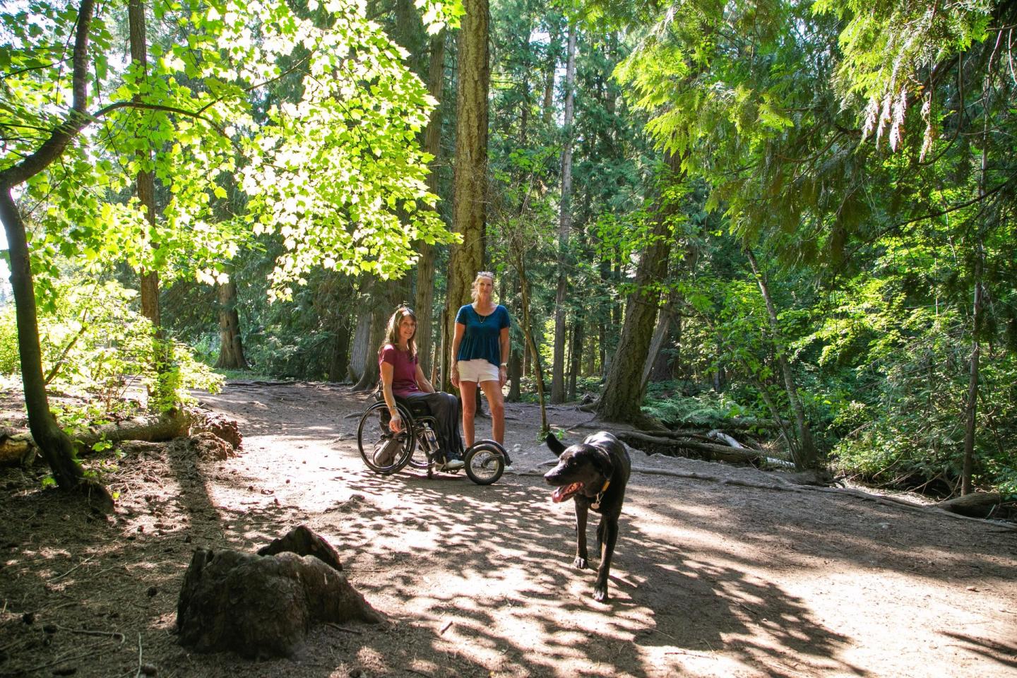Two women and a dog enjoying a sunny forest trail.