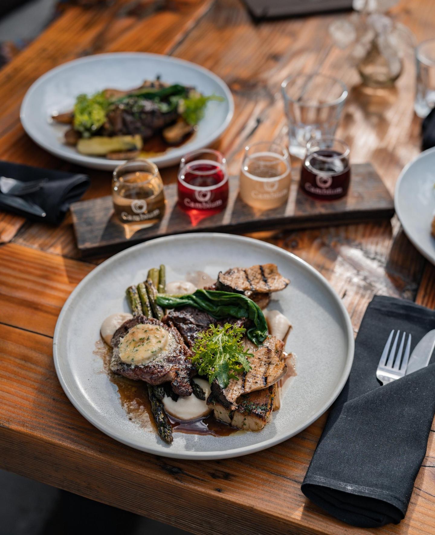 Gourmet meal with vegetables and steak on wooden table, flight of drinks in background.
