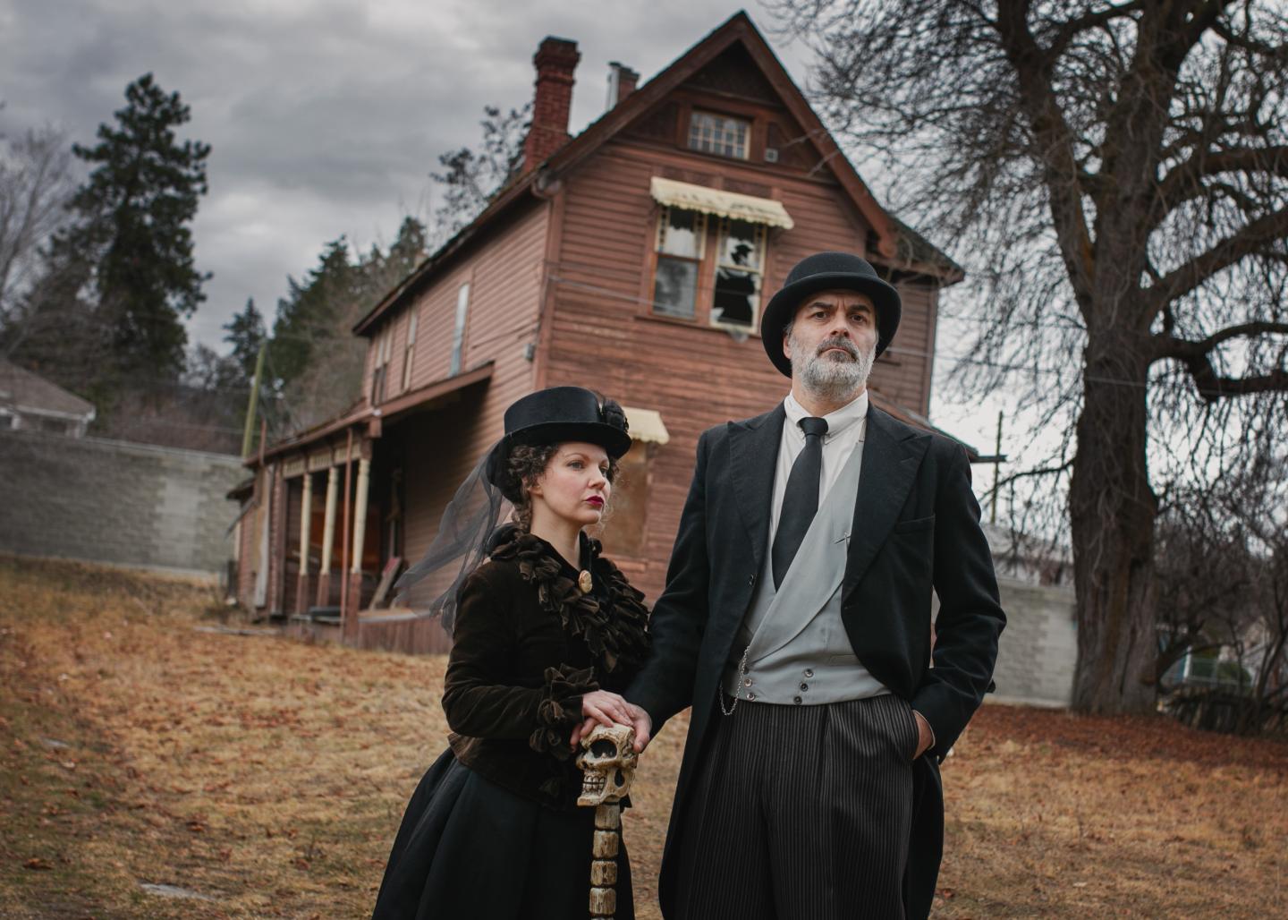 Couple in vintage clothing stand outside a Victorian house on a cloudy day.