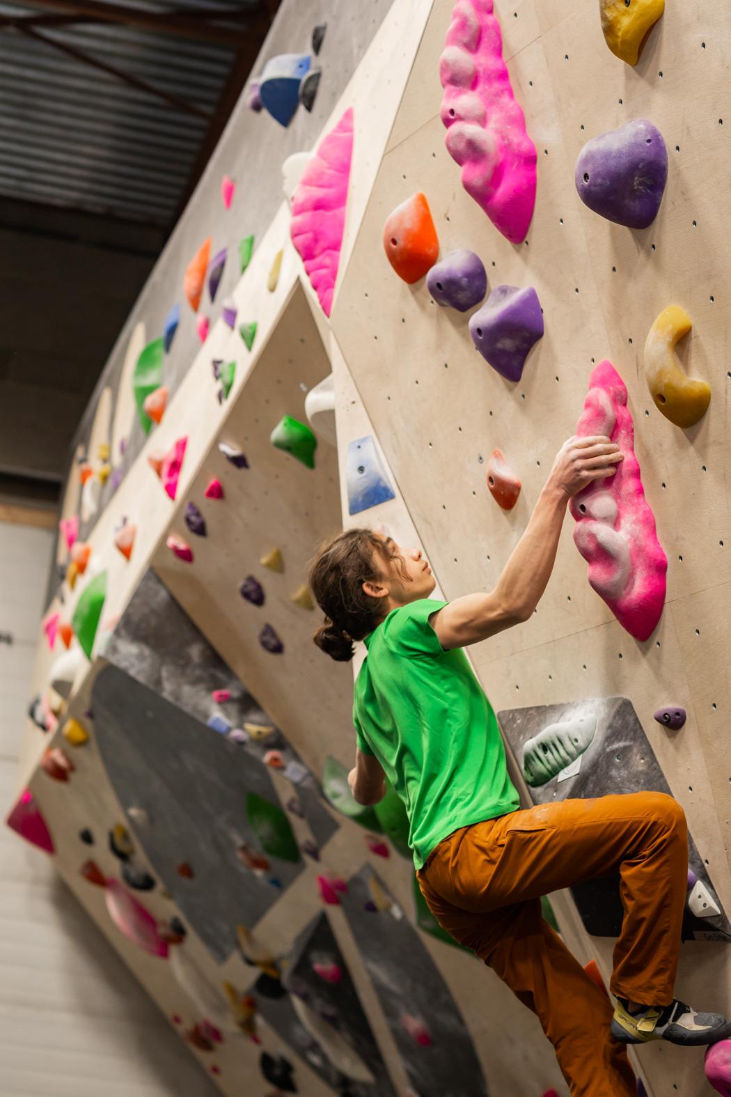 Climber in green shirt scales indoor rock wall with colorful holds.