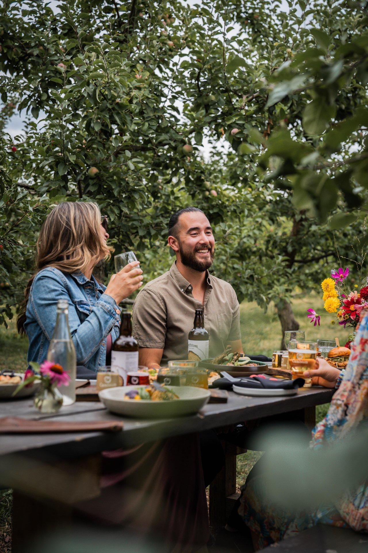 People dining outdoors, enjoying food among greenery, smiling and chatting.