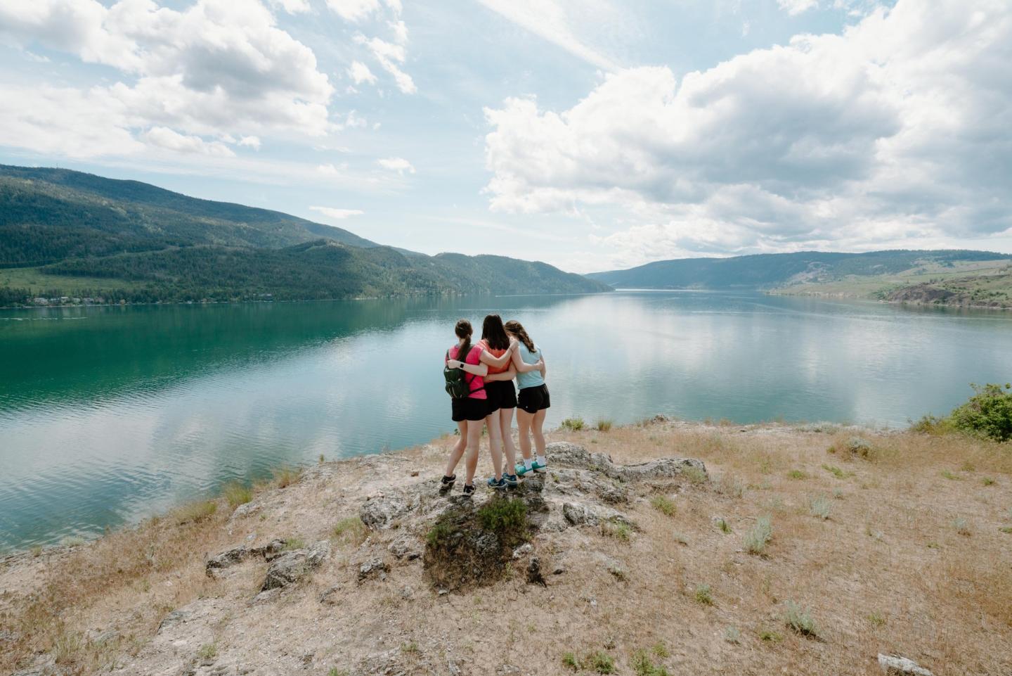 Three people embracing on a hill overlooking a lake and mountains under a cloudy sky.