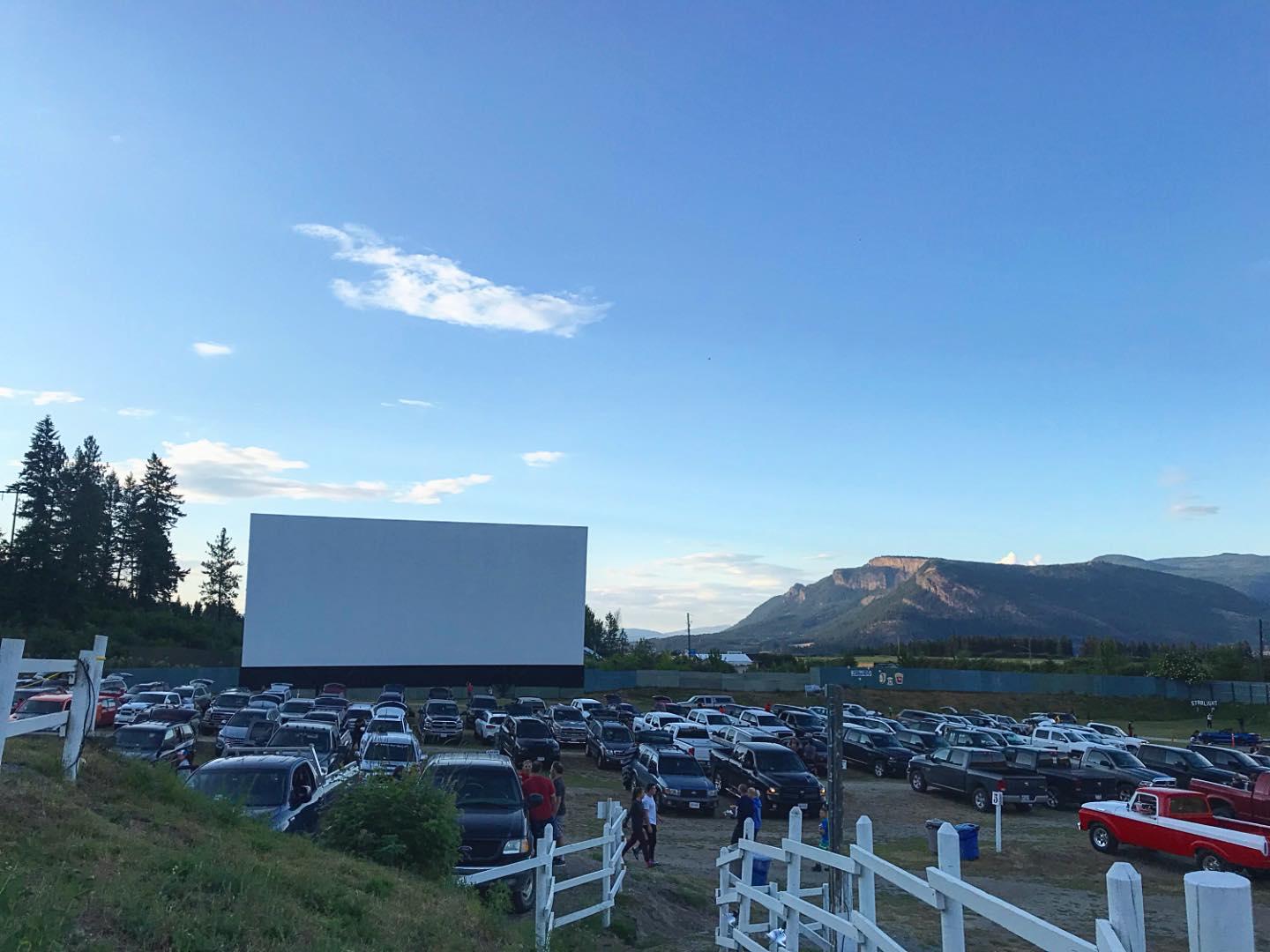 Outdoor drive-in theater with cars parked, mountains in the background under a blue sky.