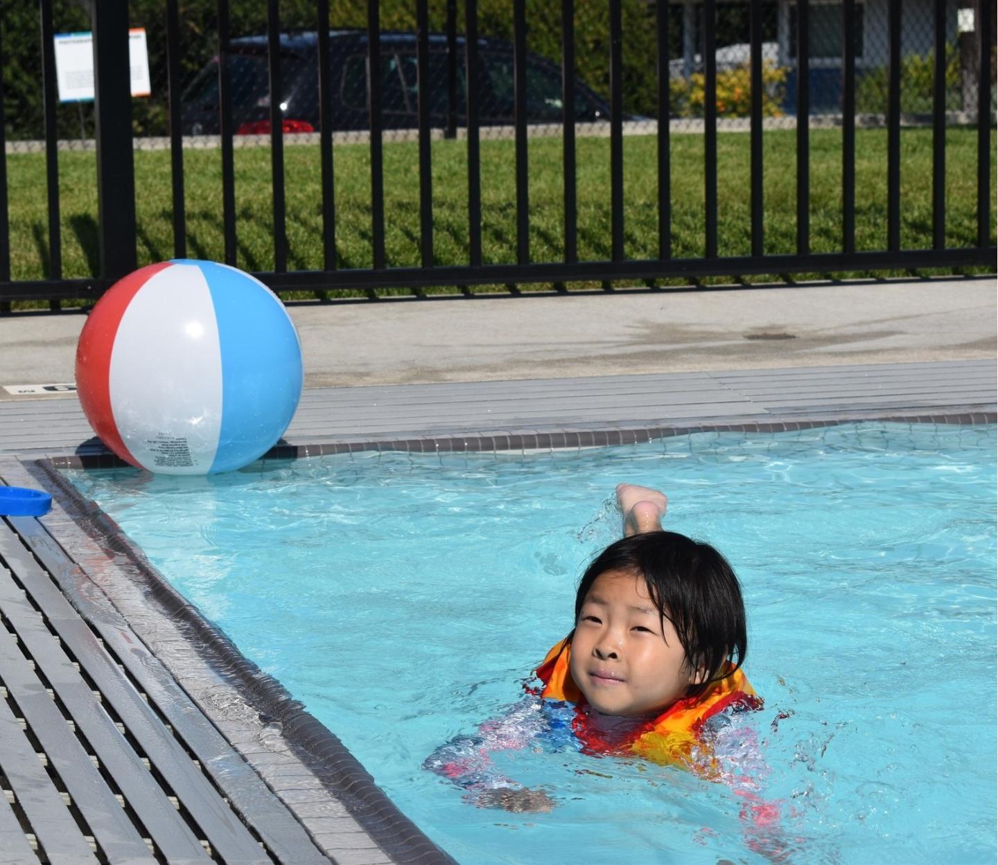 Child in pool with a life vest, beach ball on the edge, sunny day.
