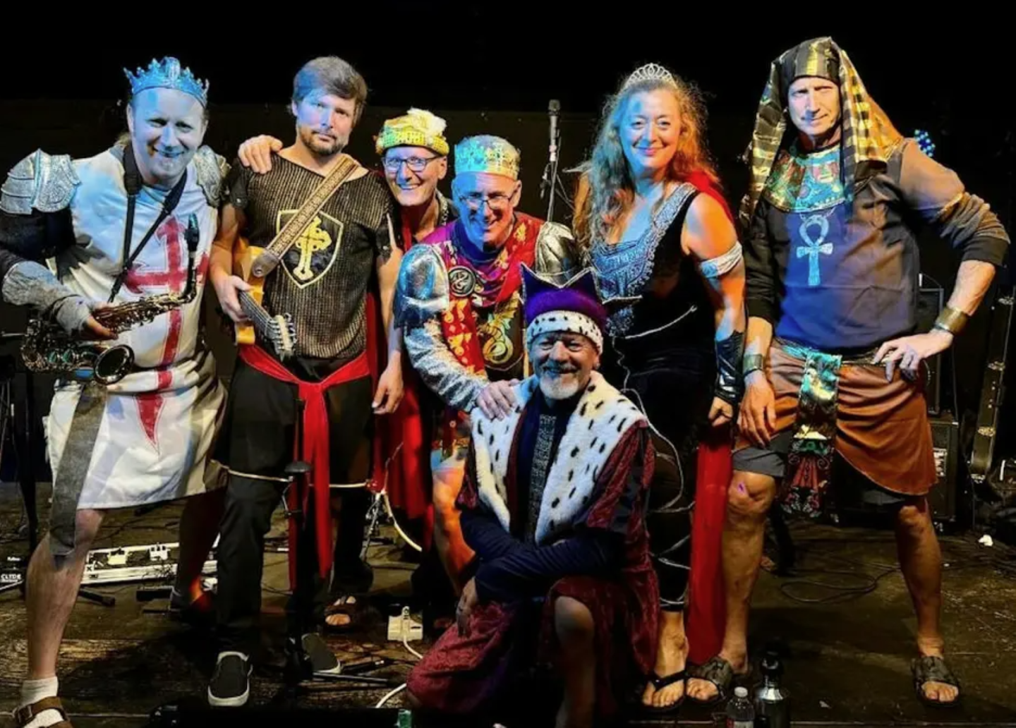 Seven people in colorful medieval and Egyptian costumes smiling on stage.