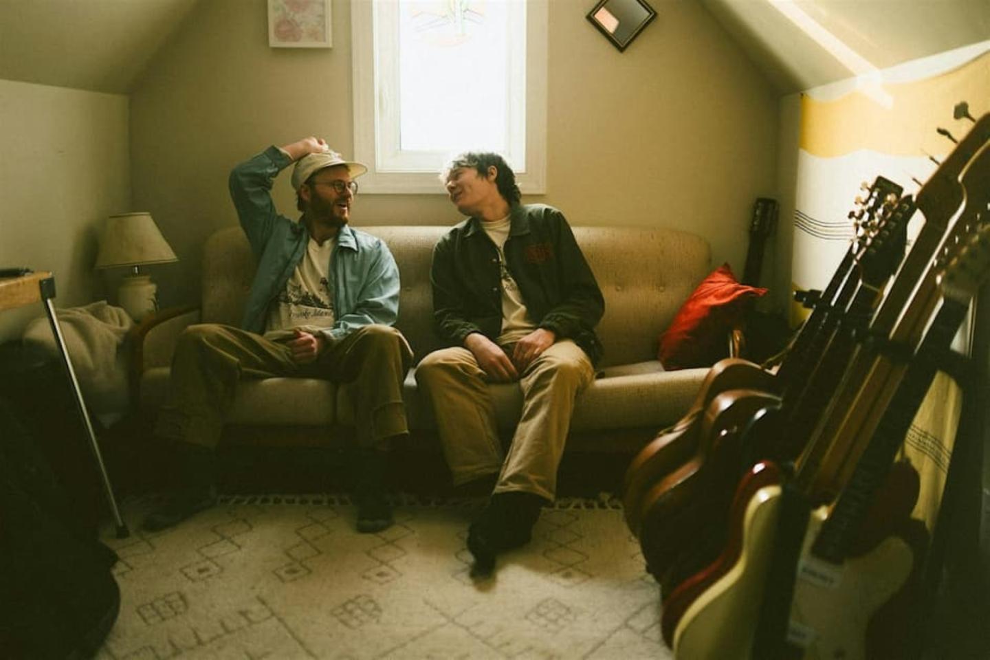 Two people sitting on a sofa in a cozy room with guitars nearby.
