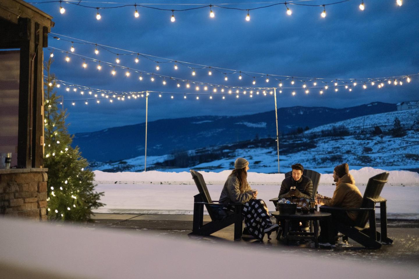 Three people dining outdoors under string lights with snowy mountains behind.