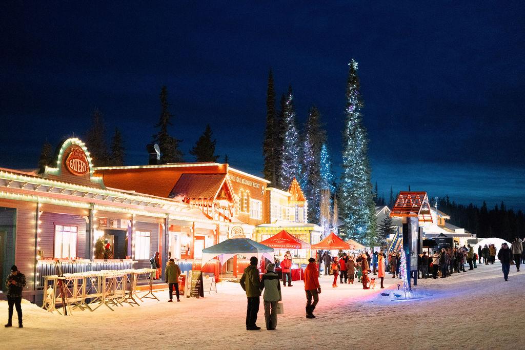 Snowy village street with warmly lit buildings and people at night.