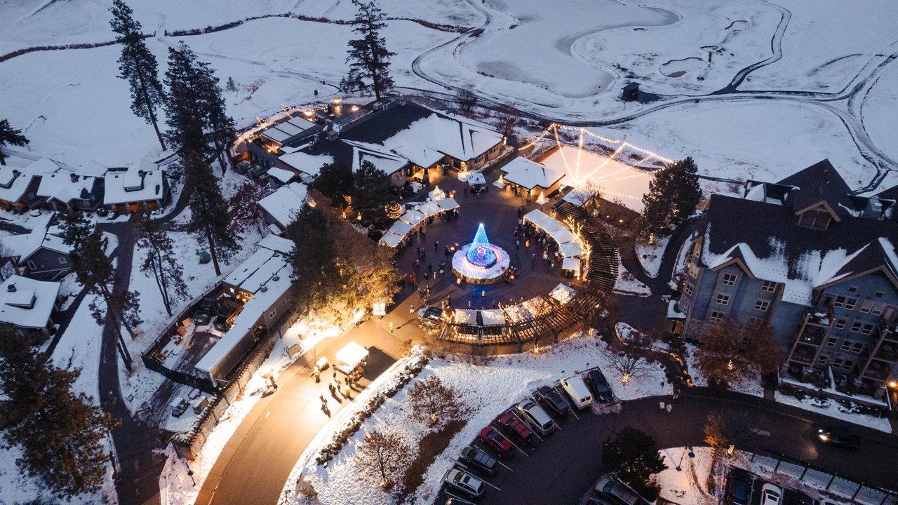 Aerial view of a snowy village square with a lit Christmas tree at dusk.